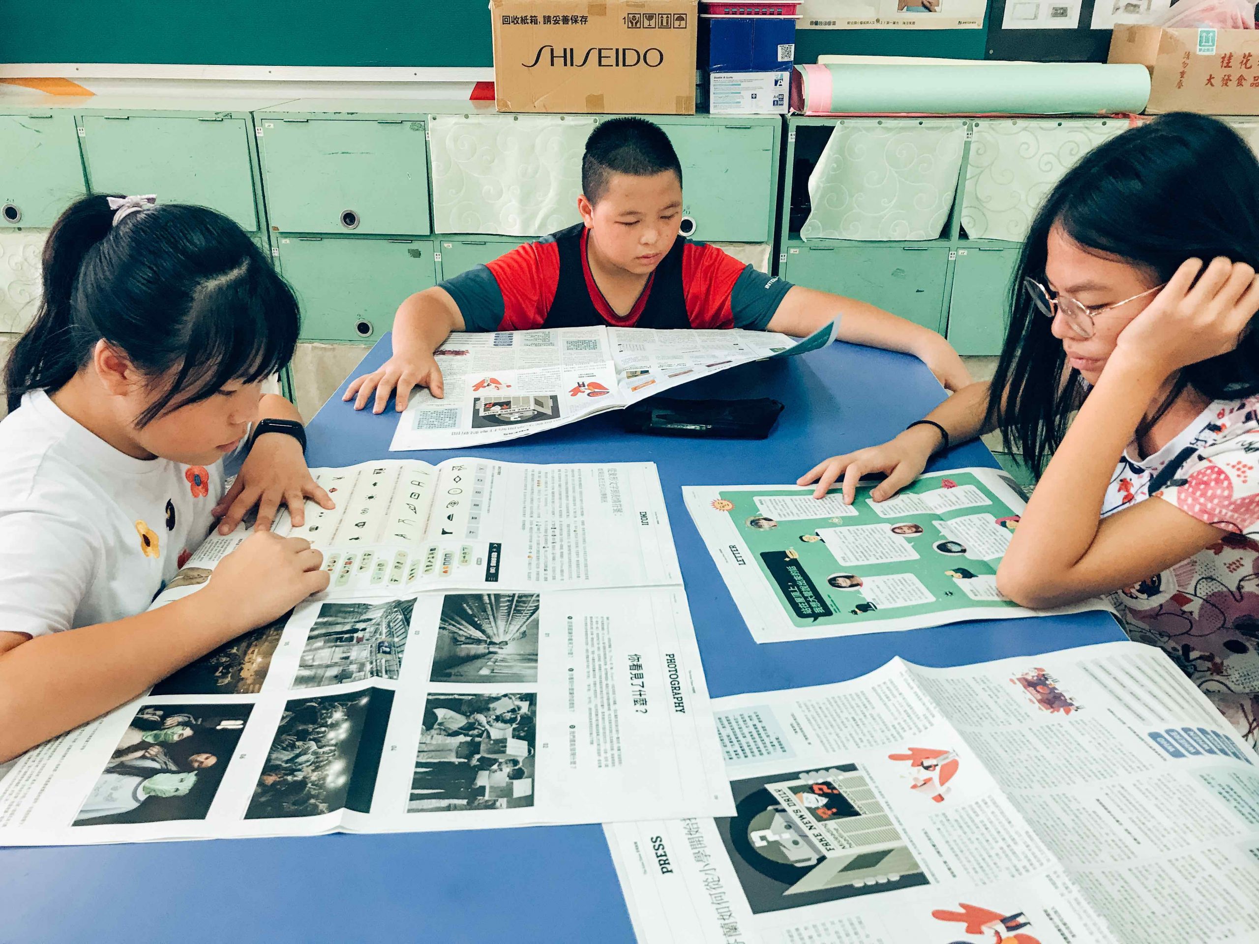 兩名女同學以及一名男同學在藍色桌子上閱讀安妮新聞報紙。